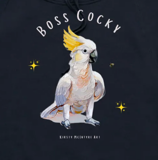 Black hoodie with a cockatoo illustration and 'Boss Cocky' text on a dark background. Close up of a a navy blue t-shirt with a parrot graphic and text 'Boss Cocky' on a white background. Original cockatoo tshirt design by artist, Kirsty McIntyre, australian.