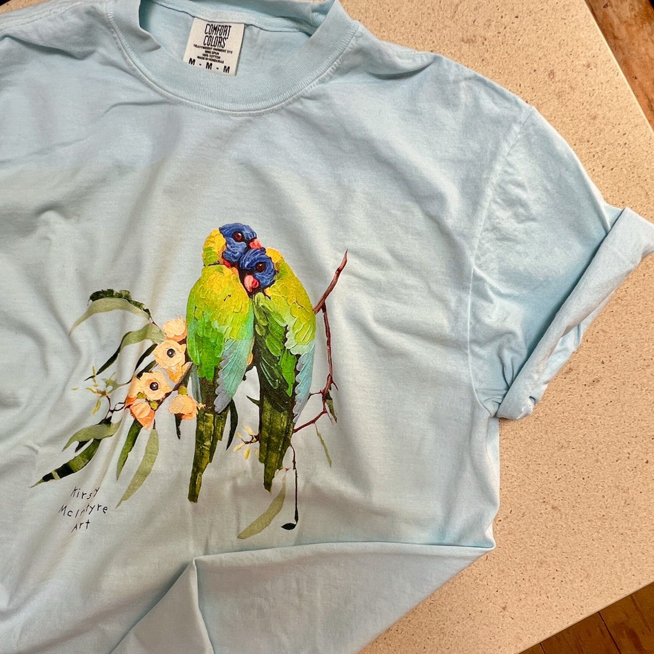 Light blue  comfort colours t-shirt with a colorful parrot design on a beige surface featuring original rainbow lorikeets cuddling on gum blossom graphic design by Australian artist, Kirsty McIntyre