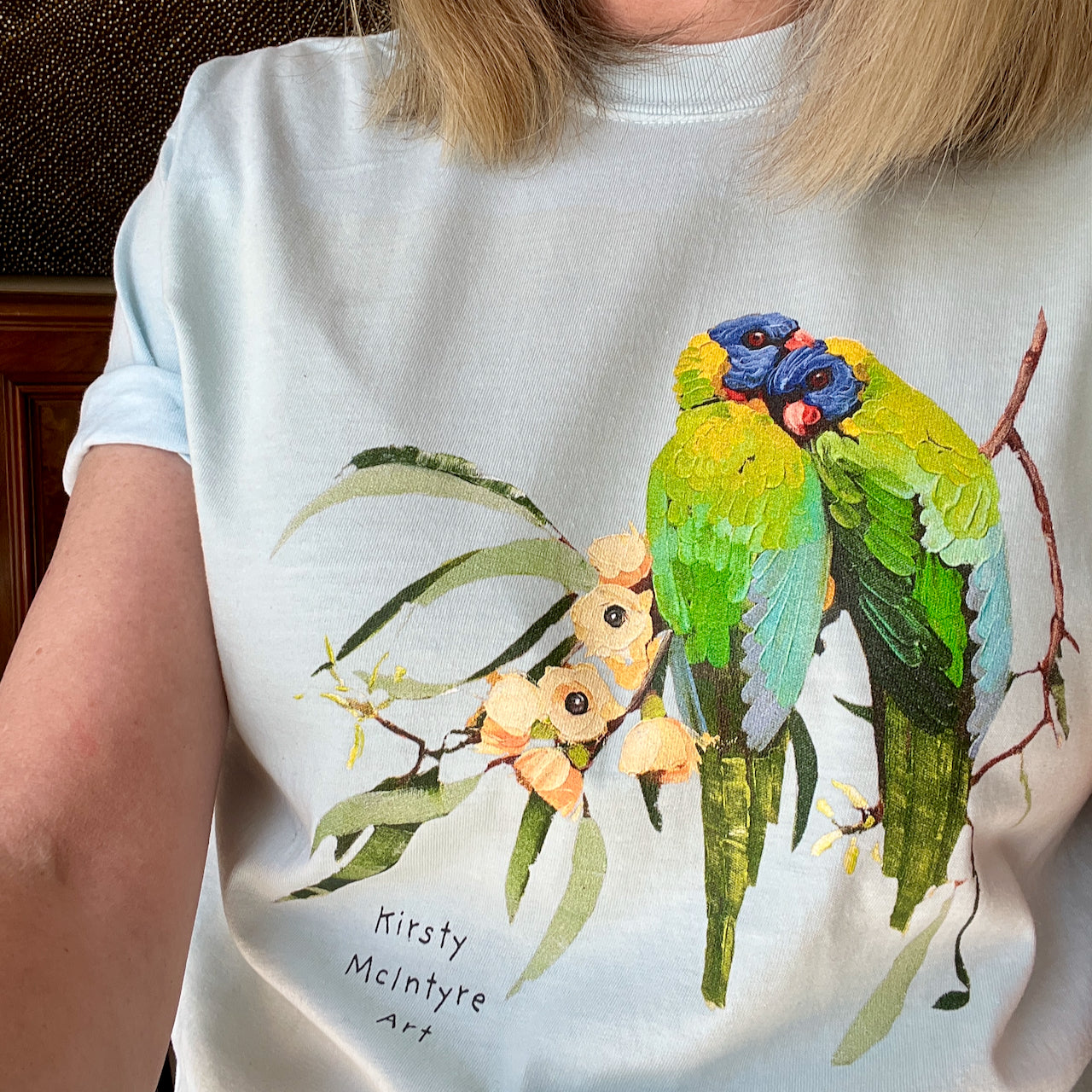 Person wearing a pale bluet-shirt with a colorful parrot design and 'Kirsty McIntyre Art' text. featuring original rainbow lorikeets cuddling on gum blossom graphic design by Australian artist, Kirsty McIntyre