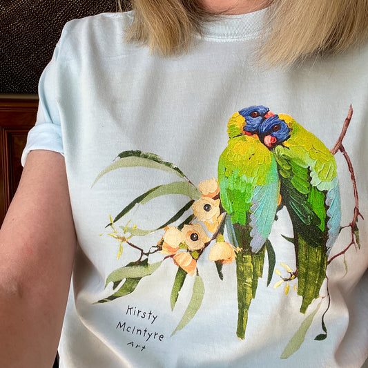 Person wearing a pale bluet-shirt with a colorful parrot design and 'Kirsty McIntyre Art' text. featuring original rainbow lorikeets cuddling on gum blossom graphic design by Australian artist, Kirsty McIntyre