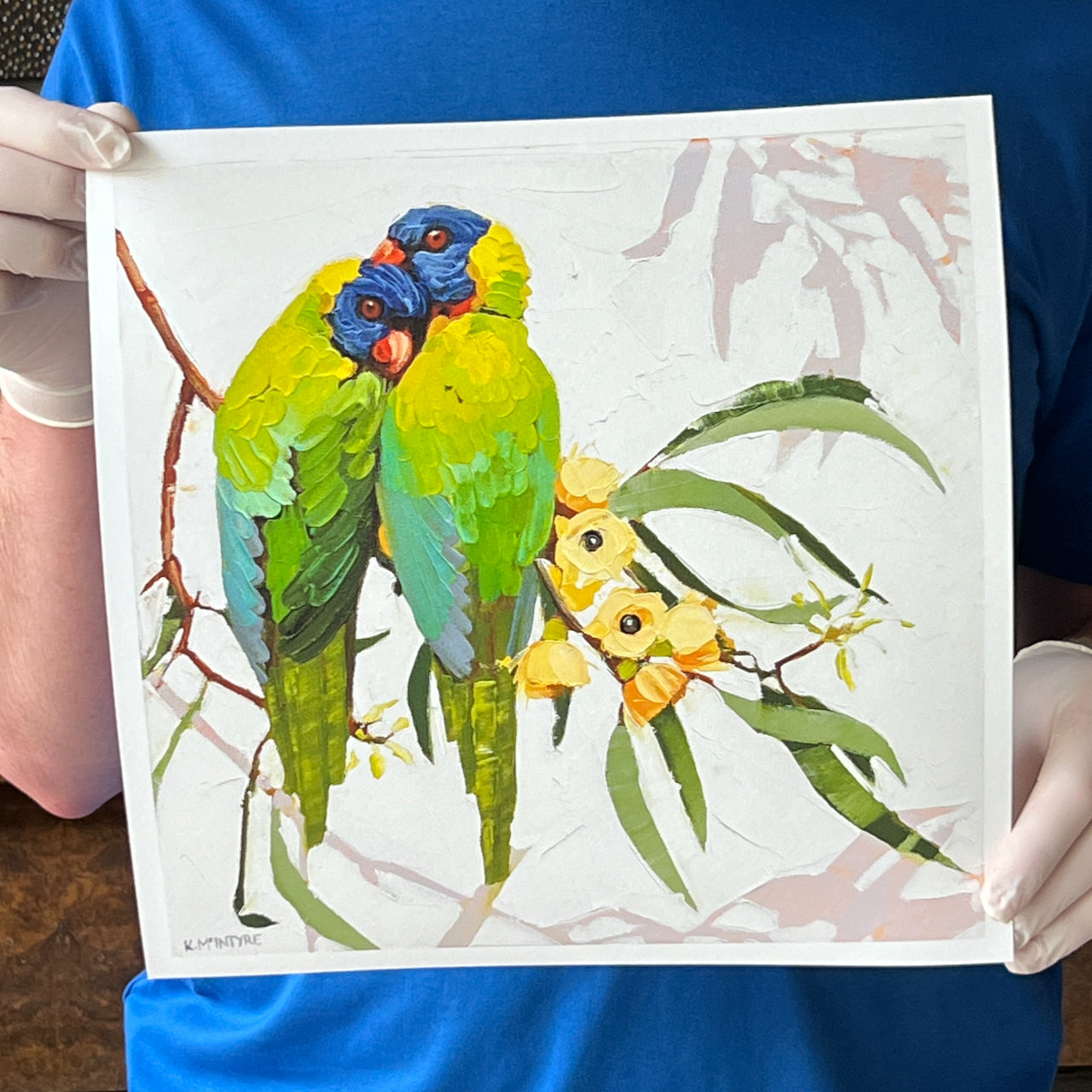 Person holding a painting of two parrots on a branch with leaves and flowers. Original rainbow lorikeet design by Australian artist, Kirsty McIntyre available as a giclee reproduction print