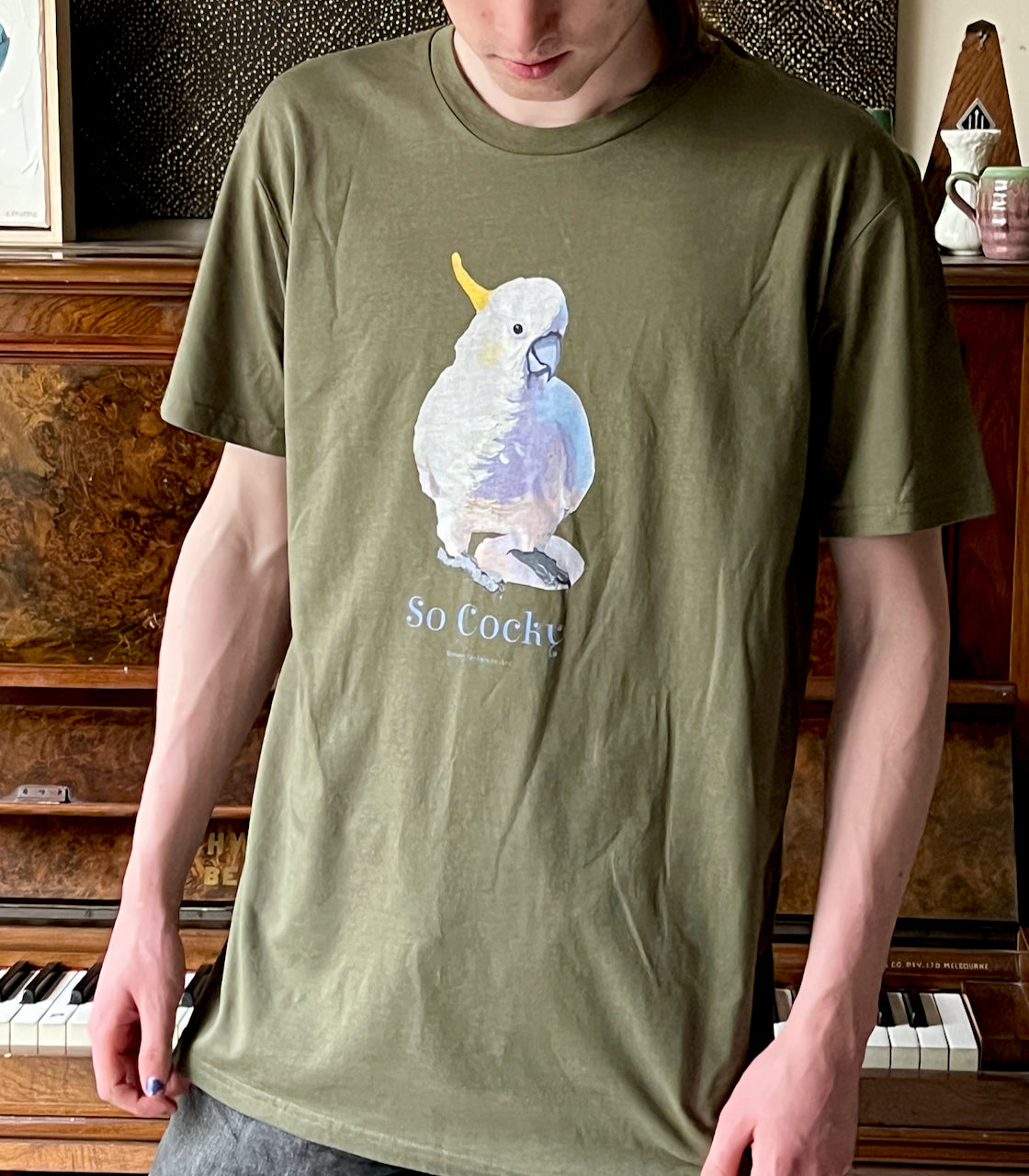 Person wearing a green t-shirt with a white cockatoo parrot graphic and 'So Cocky' text. Original tshirt design by best popular australian bird artist, Kirsty McIntyre