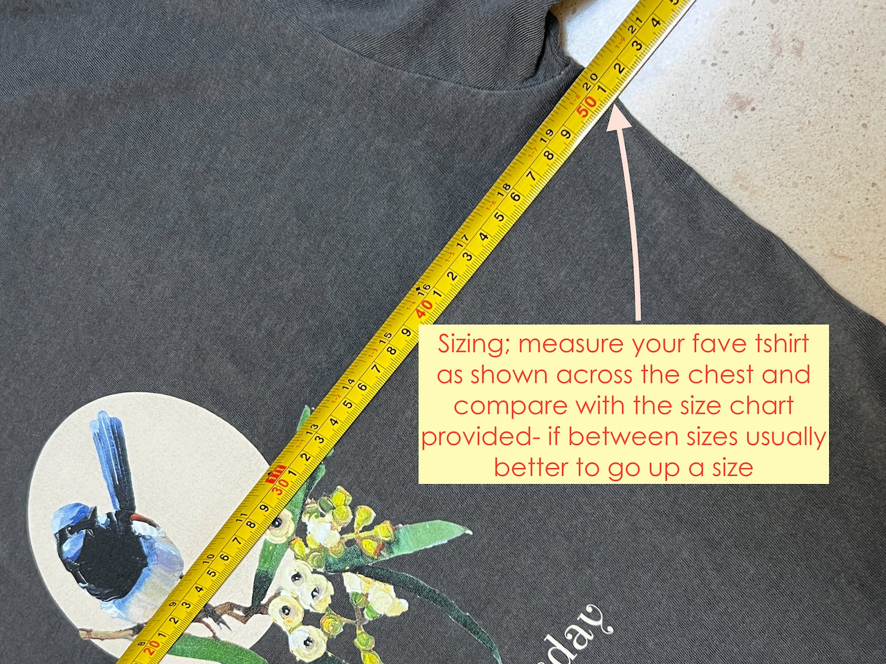 Gray t-shirt with a graphic design and a measuring tape across it, with text on sizing instructions. tsirt withsuperb blue wren bird design and text 'Every day is Wrensday' on a branch with leaves and flowers. original design by artist australian Kirsty McIntyre- demonstrates measuring the hoodie for sizing