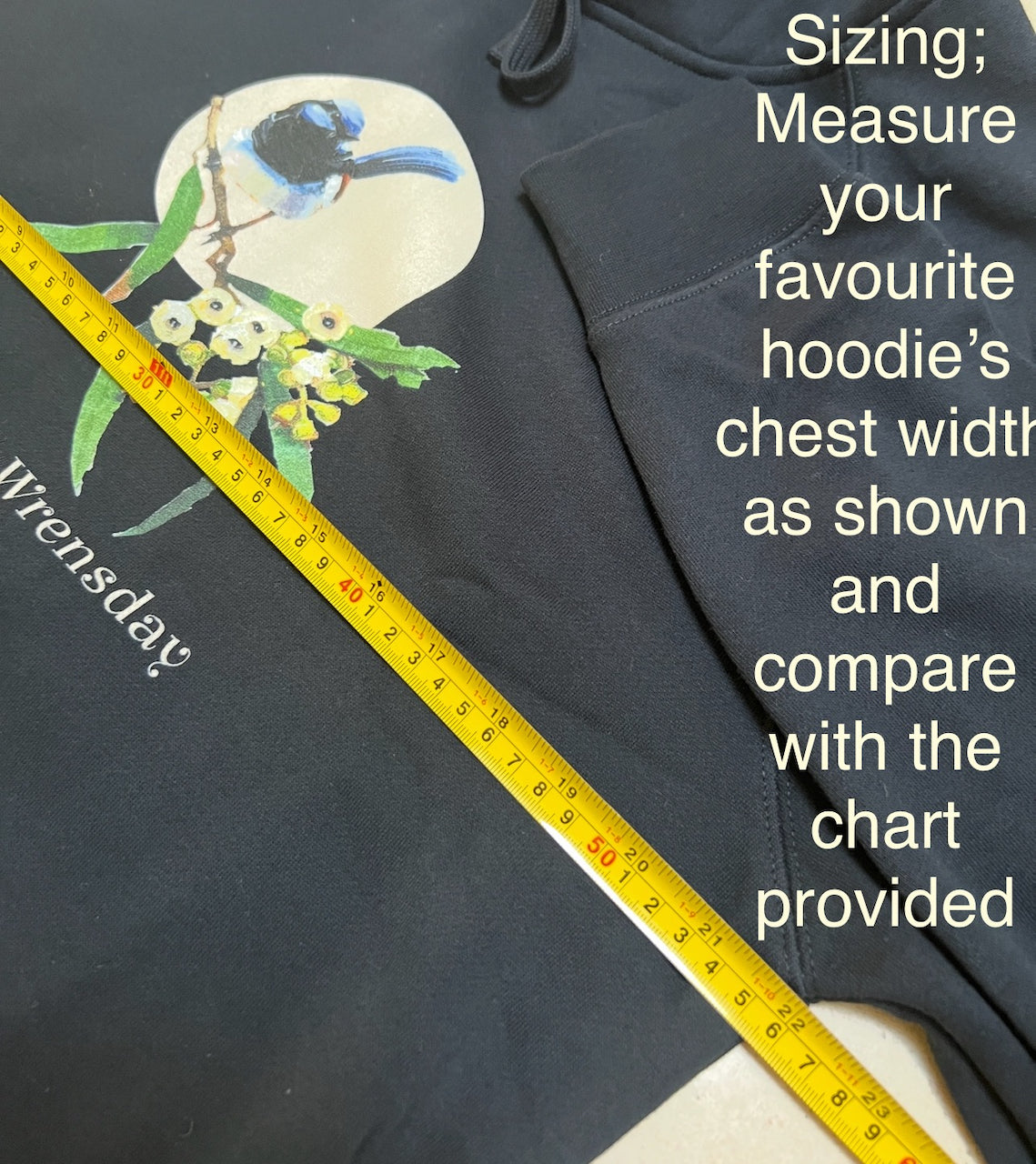 person wearing a navy blue hoodie with a superb blue wren bird design and text 'Every day is Wrensday' on a branch with leaves and flowers. original design by artist australian Kirsty McIntyre- demonstrates measuring the hoodie for sizing